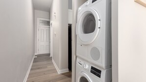 Une buanderie avec des machines à laver et des sèche-linge superposés, un plancher en bois et une porte blanche au fond du couloir.