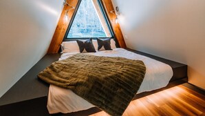 Une pièce triangulaire comprenant un lit, un plancher en bois et une fenêtre donnant sur des arbres.