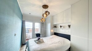A modern bedroom with a large bed, a ceiling light fixture, and a view of the outdoors through a window. - Flat Las Vegas - Seaview (Middelkerke)