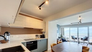 A modern kitchen with a built-in dishwasher, a dining area with a round table and chairs, and a view of the ocean through large windows. - Flat Las Vegas - Seaview (Middelkerke)