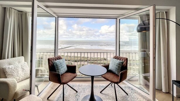 A room with a large window offering a view of the beach and ocean. There is a round table with two chairs, a sofa, and a floor lamp. - Flat Las Vegas - Seaview (Middelkerke)