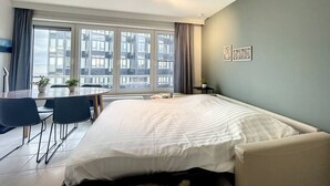 A modern hotel room with a large bed, a small desk, and a view of the city through large windows. - Flat Vendome - Studio with seaview (Middelkerke)