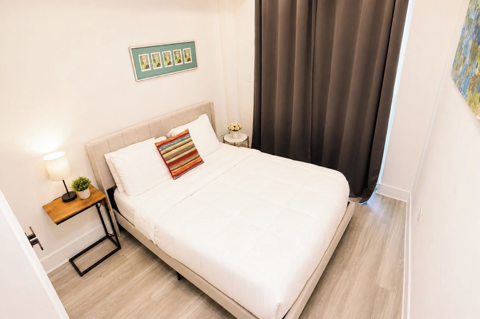 A neatly made bed with a striped pillow, a bedside table with a lamp and a small plant, and a framed picture on the wall.