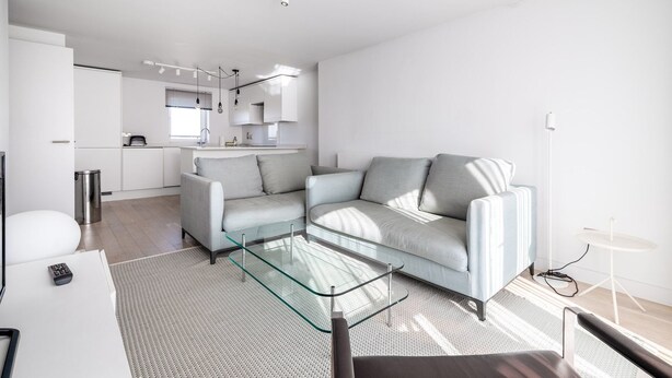 A modern living room with a glass coffee table, a grey sofa, and a kitchen area in the background.