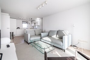 A modern living room with a glass coffee table, a grey sofa, and a kitchen area in the background.