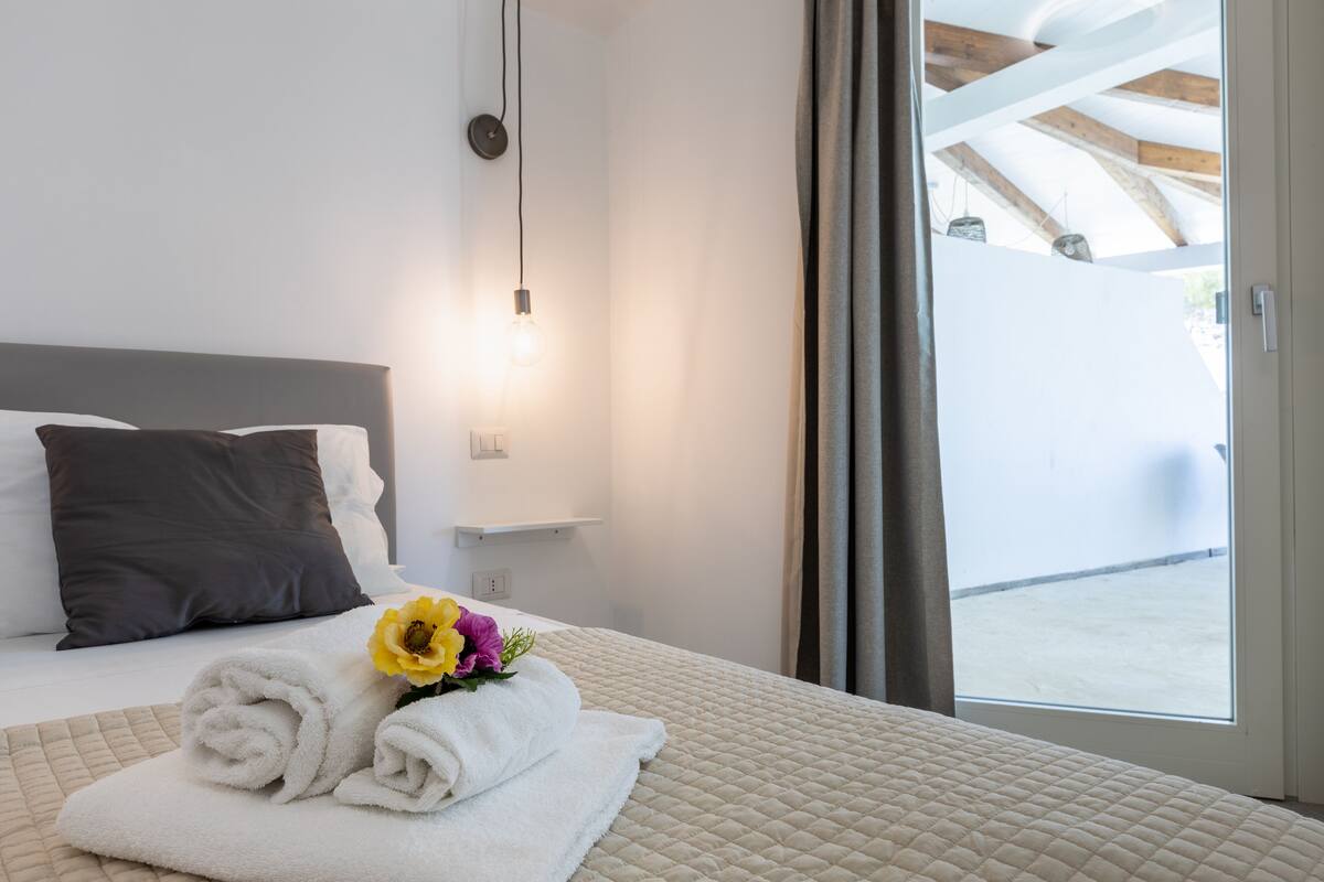A hotel room with a bed, a nightstand, a window with curtains, and a view of the outside.