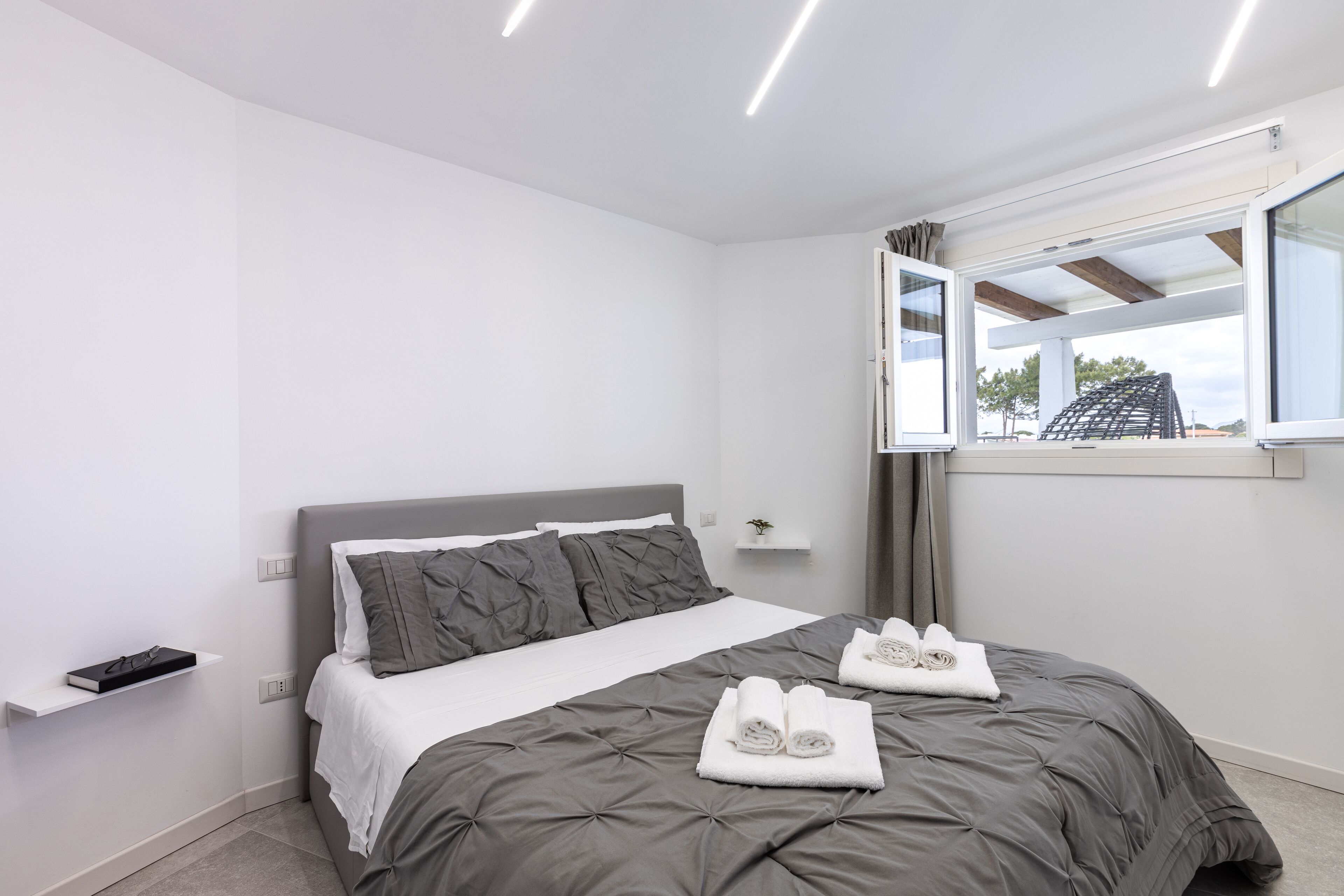 A modern bedroom with a grey bed, white walls, and a window with curtains.