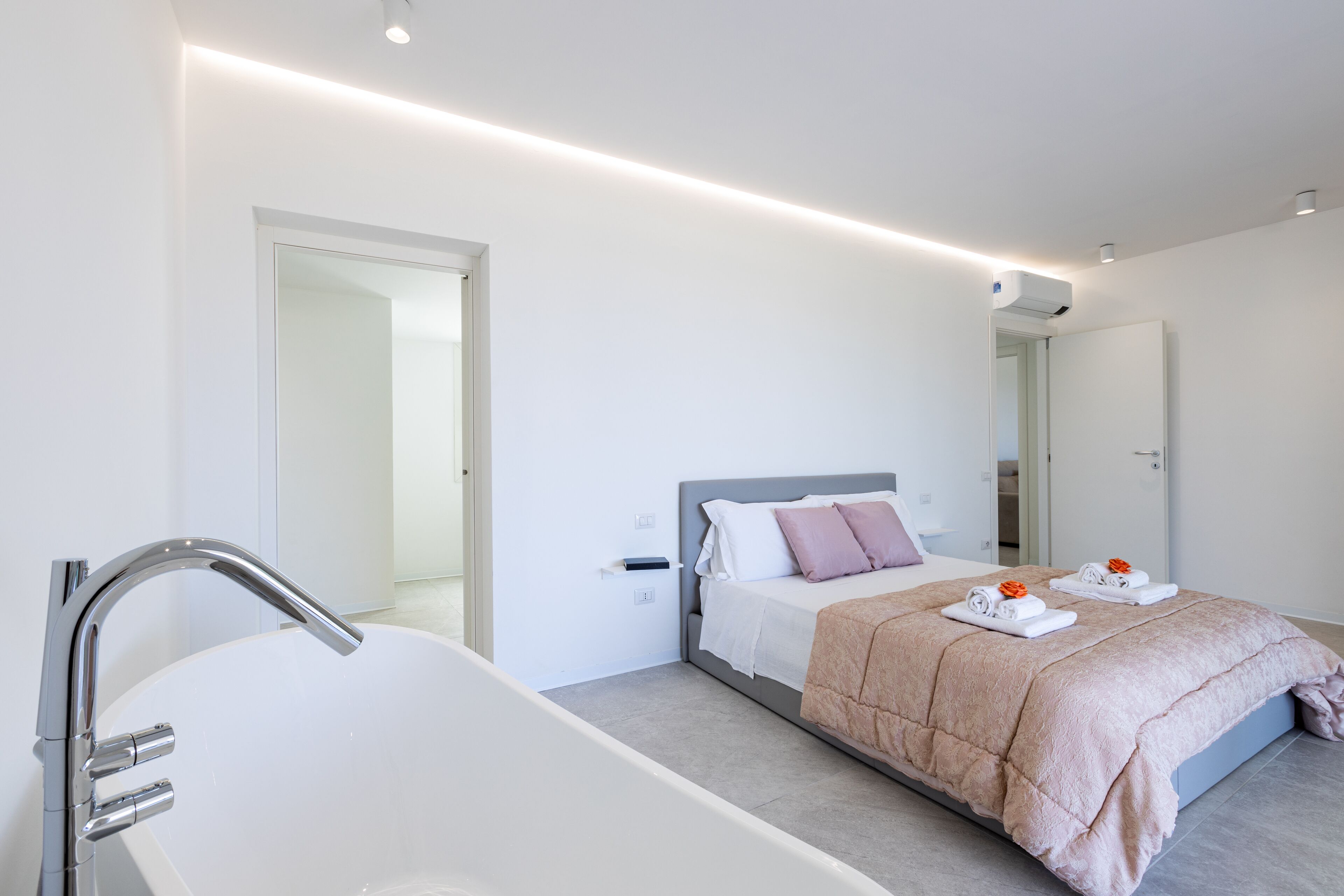Et moderne hotelværelse med et stort, hvidt badekar, en pænt redt seng med et pink tæppe og et stort spejl på væggen.