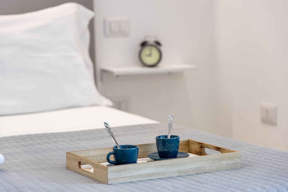 A bed with a white pillow and a wooden tray with two blue cups and a spoon on a quilted bedspread.