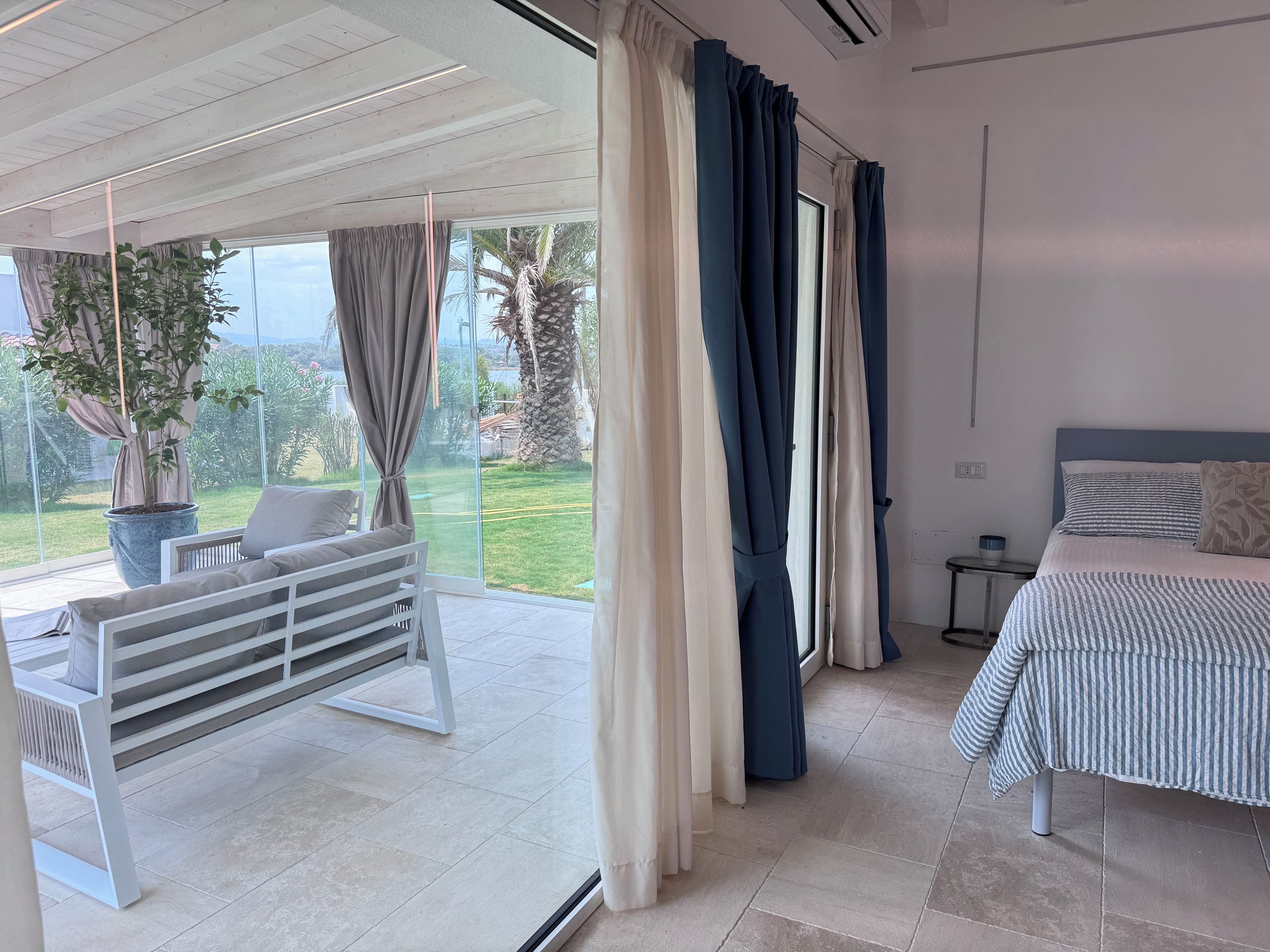 A bedroom with a bed, a bedside table, a window with curtains, and a view of a garden.