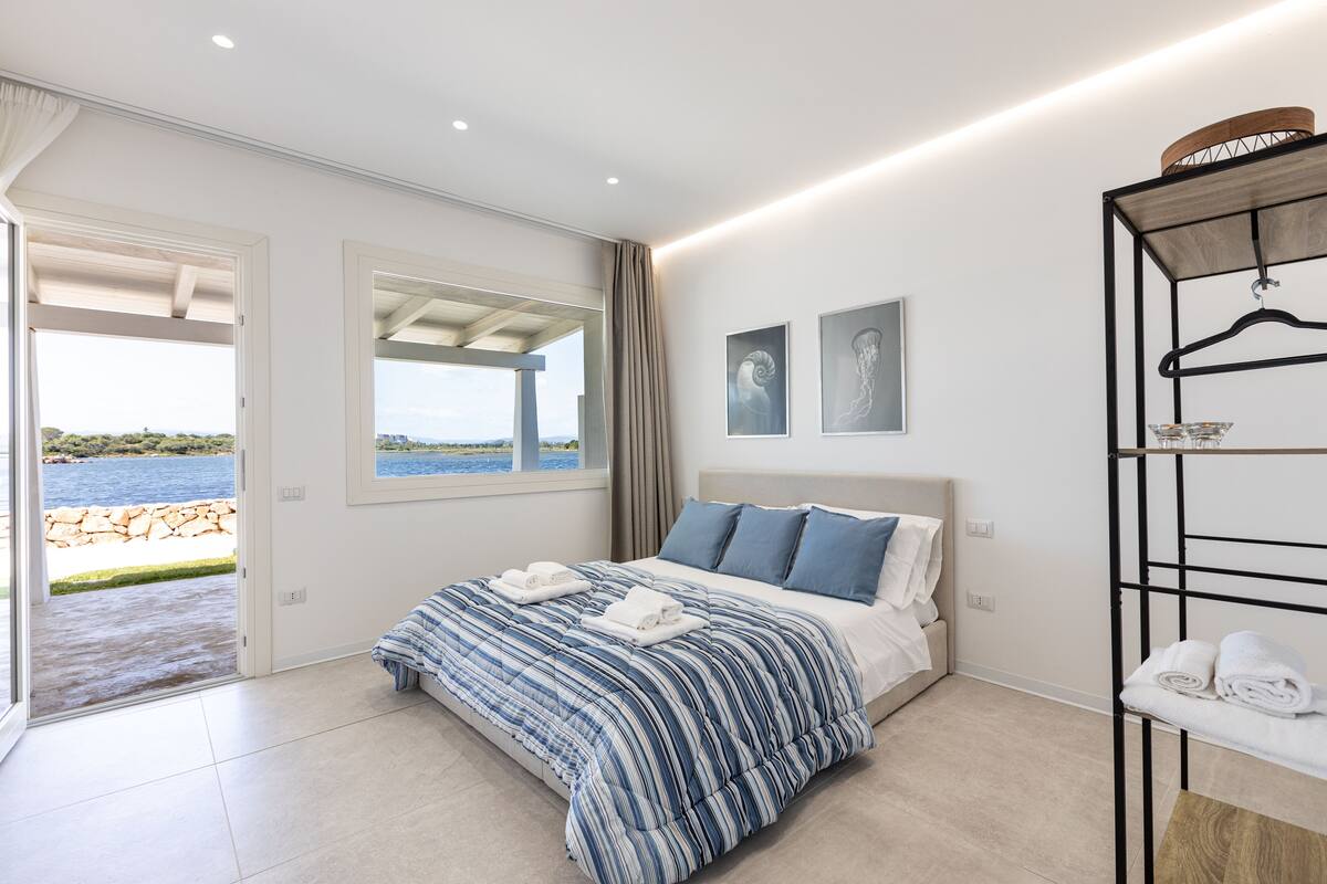 A bedroom with a large bed, a view of the sea, and a wooden shelf with towels.