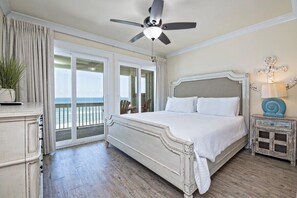 A bedroom with a large bed, a ceiling fan, a nightstand with a lamp, and a view of the ocean through a sliding door.
