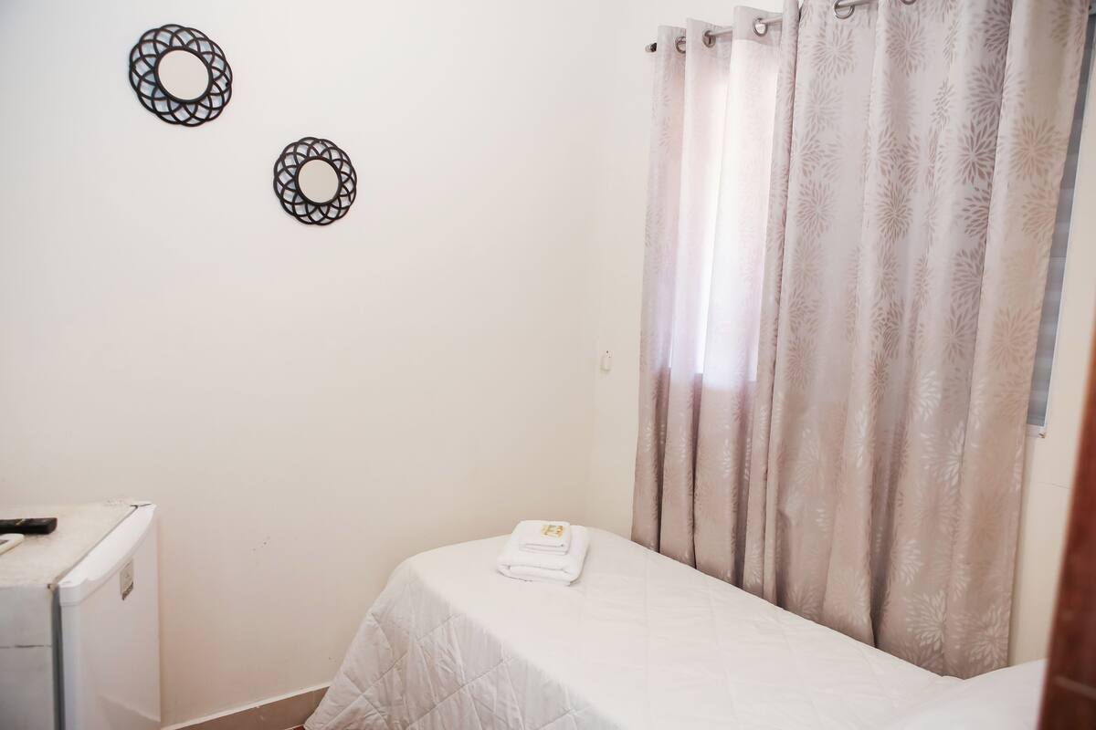Um quarto pequeno com uma cama, uma parede branca e uma cortina.