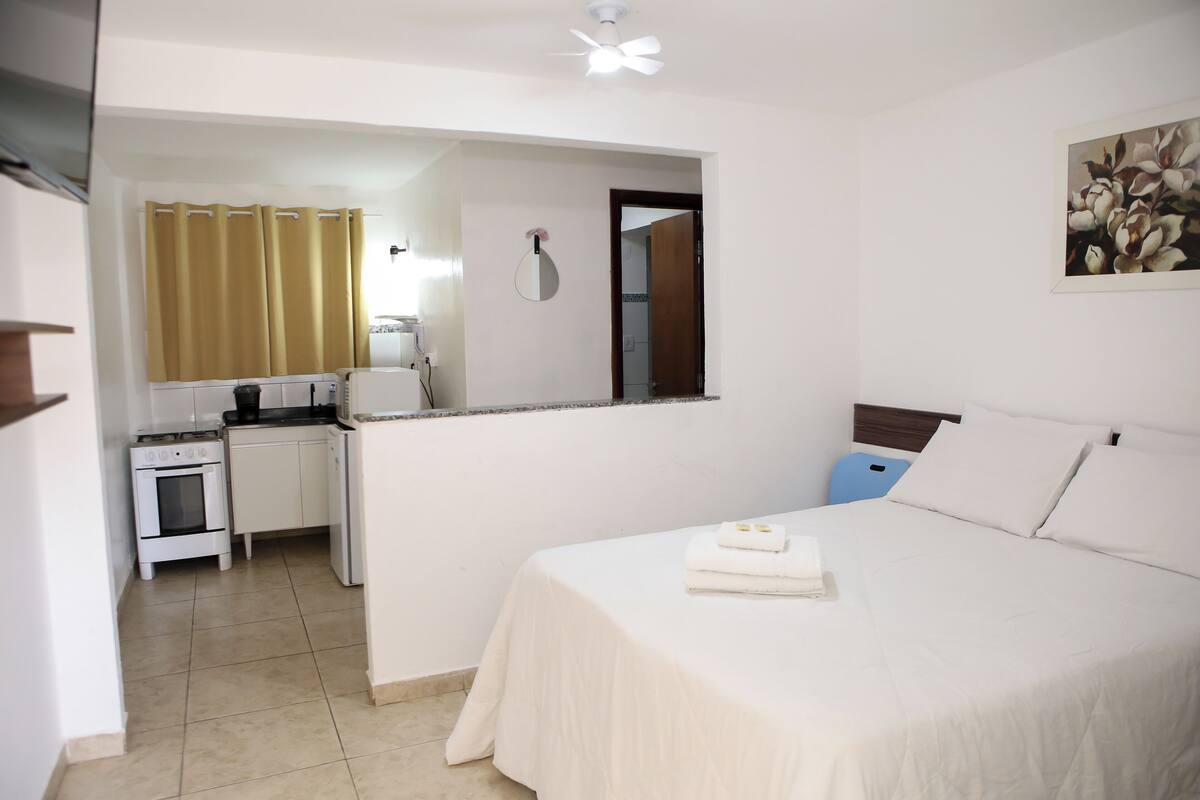 Quarto de hotel com cama, pequeno espaço de cozinha e banheiro visível por uma porta aberta.