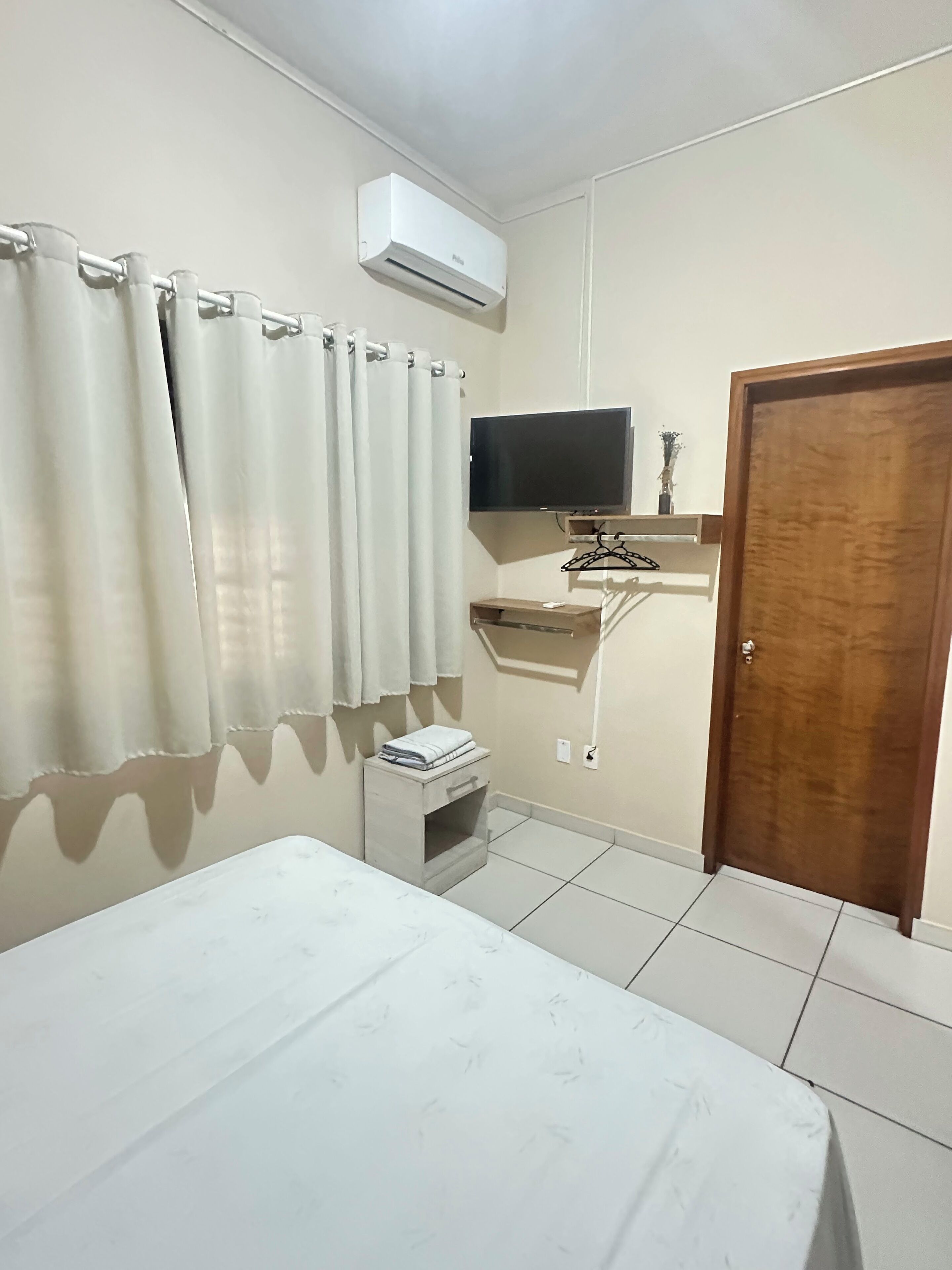 Um quarto com uma cama, uma televisão, uma porta de madeira e uma mesinha de cabeceira com um livro.