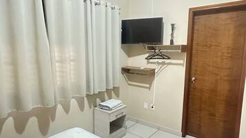 Um quarto com uma cama, uma televisão, uma porta de madeira e uma mesinha de cabeceira com um livro.