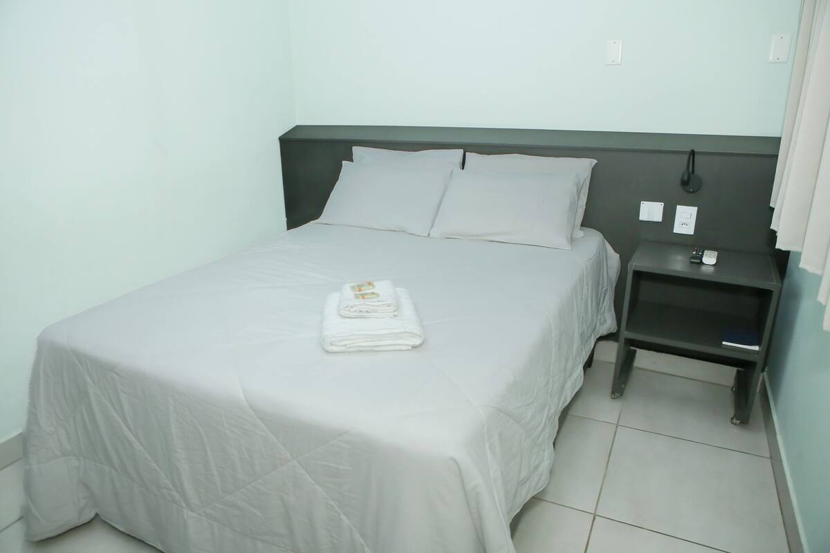 Uma cama bem arrumada, uma toalha dobrada, uma mesa de cabeceira com telefone e livro, e uma tomada na parede.