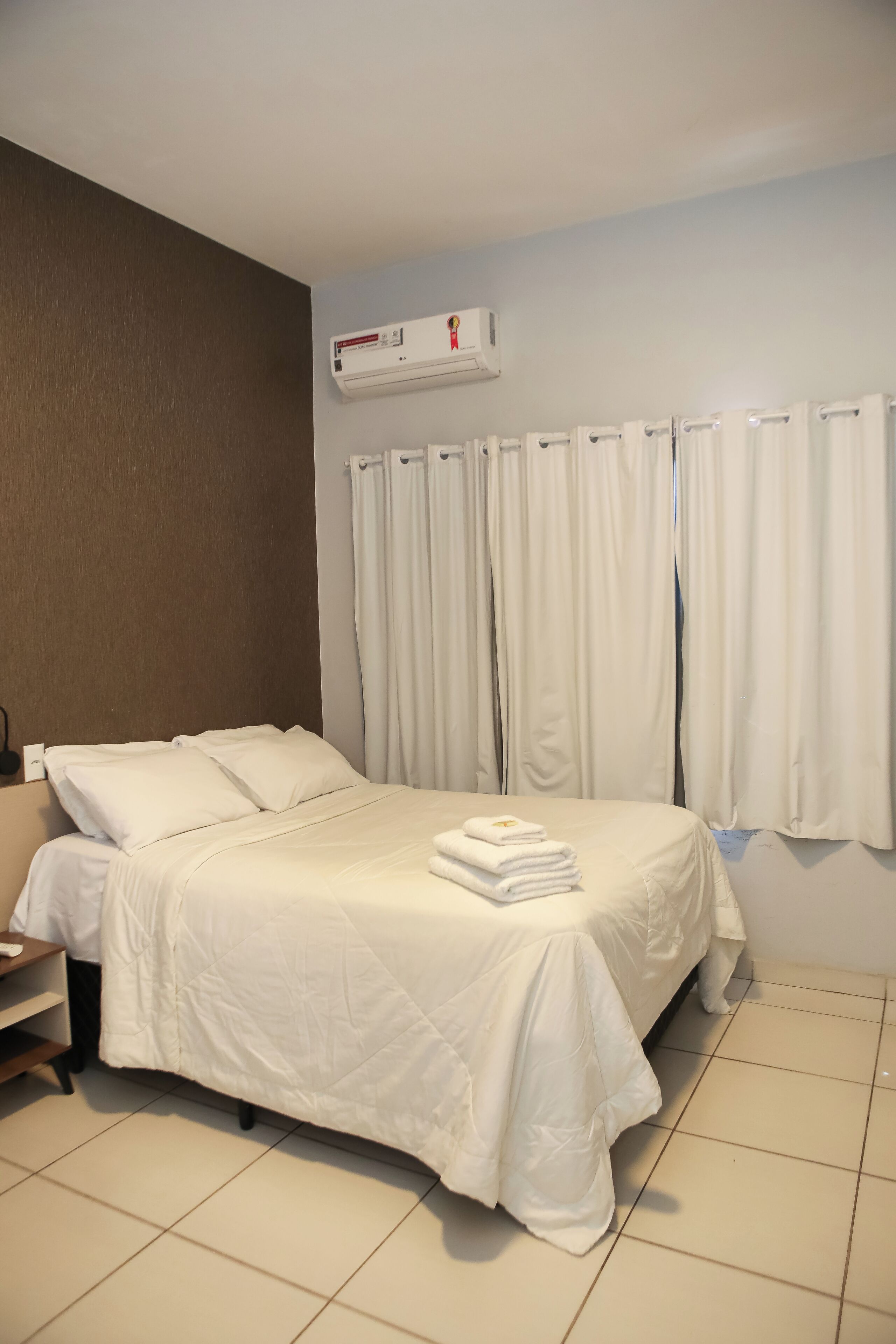 A hotel room with a bed, white curtains, a wall-mounted air conditioner, and a tiled floor.