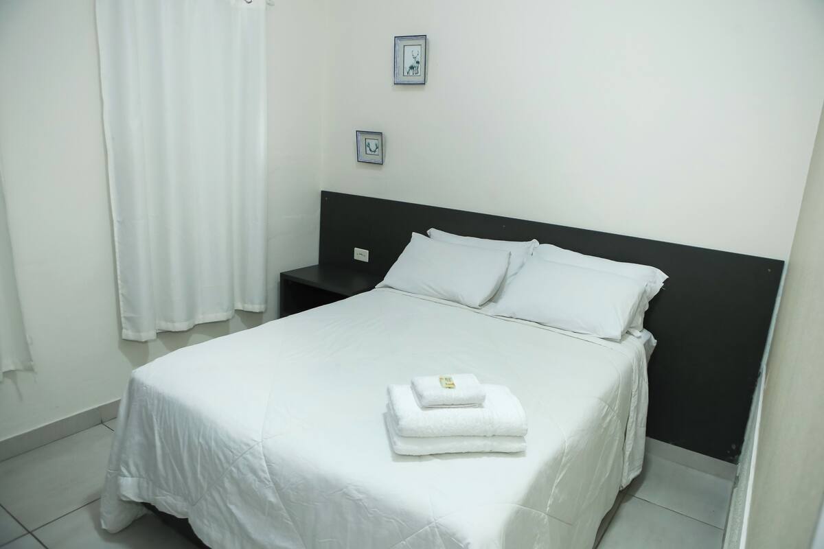 Cama bem arrumada com lençóis brancos e toalha dobrada, cabeceira preta e cortina branca.