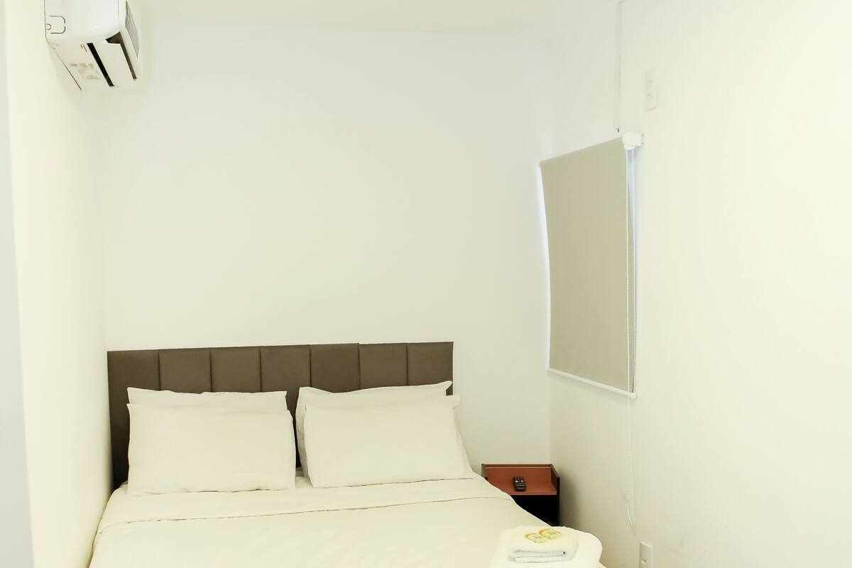 Uma cama bem arrumada com cabeceira, ar condicionado fixado na parede, mesa de cabeceira com luminária e um espelho na parede.