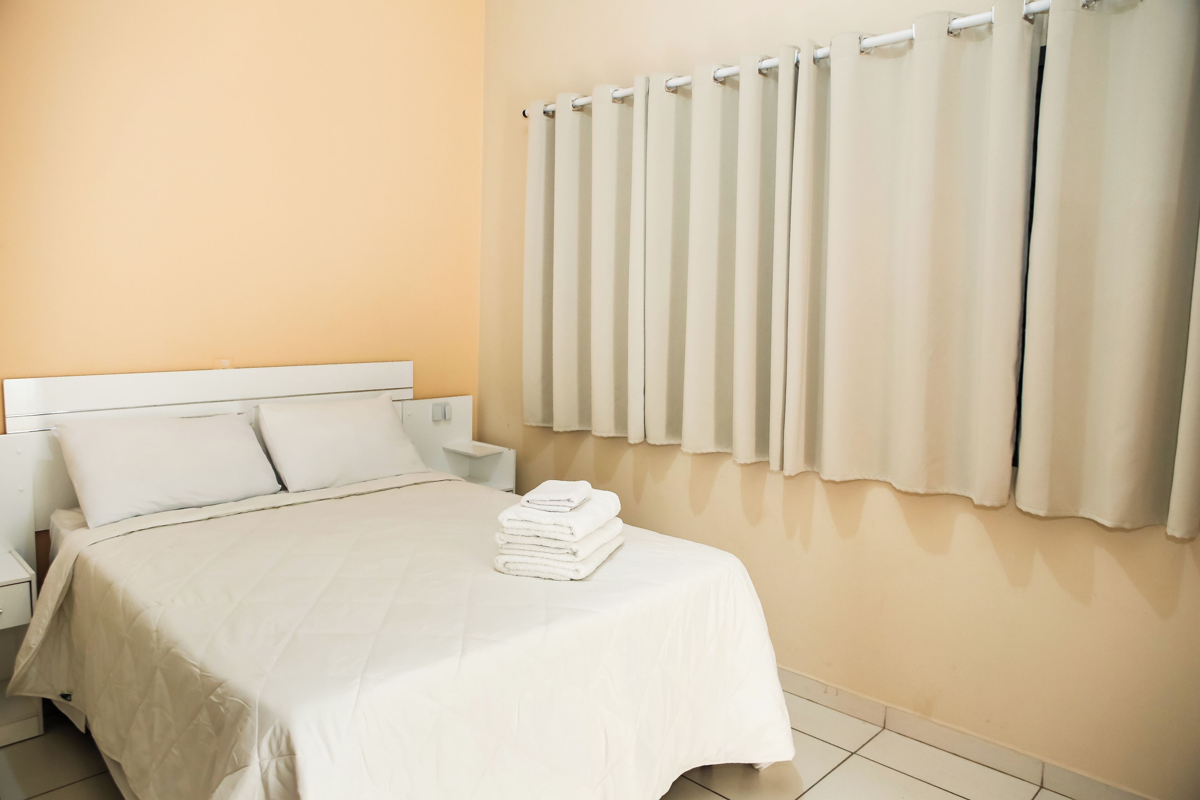 A hotel room with a bed, white curtains, a radiator, and a neatly made bed with folded towels.