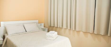 A hotel room with a bed, white curtains, a radiator, and a neatly made bed with folded towels.