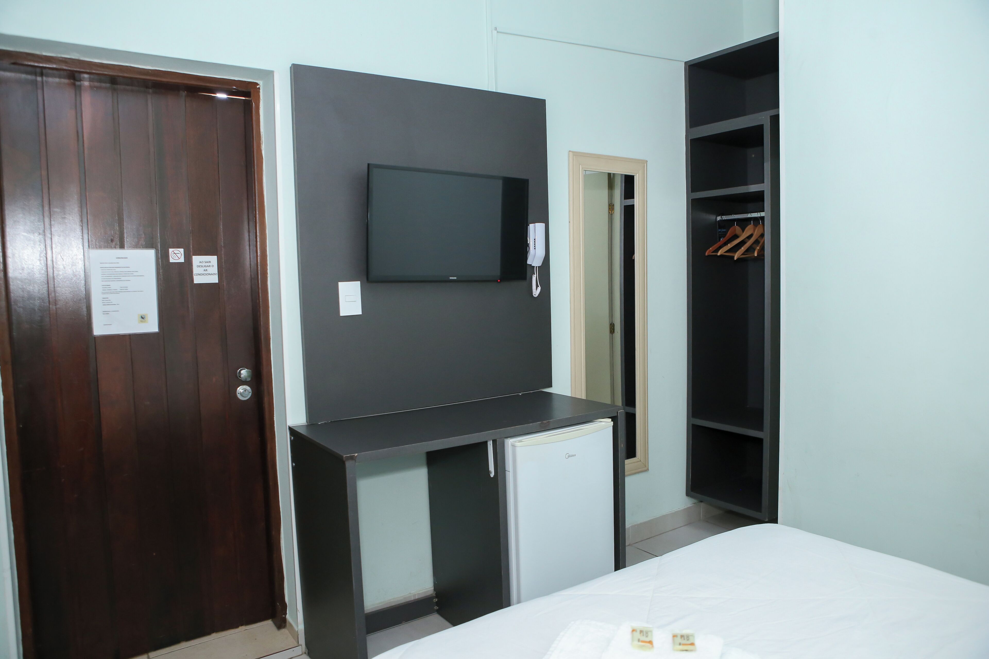 A hotel room with a wooden door, a flat-screen TV mounted on a dark wall, a small refrigerator, a closet with hanging clothes, and a bed with a white comforter.