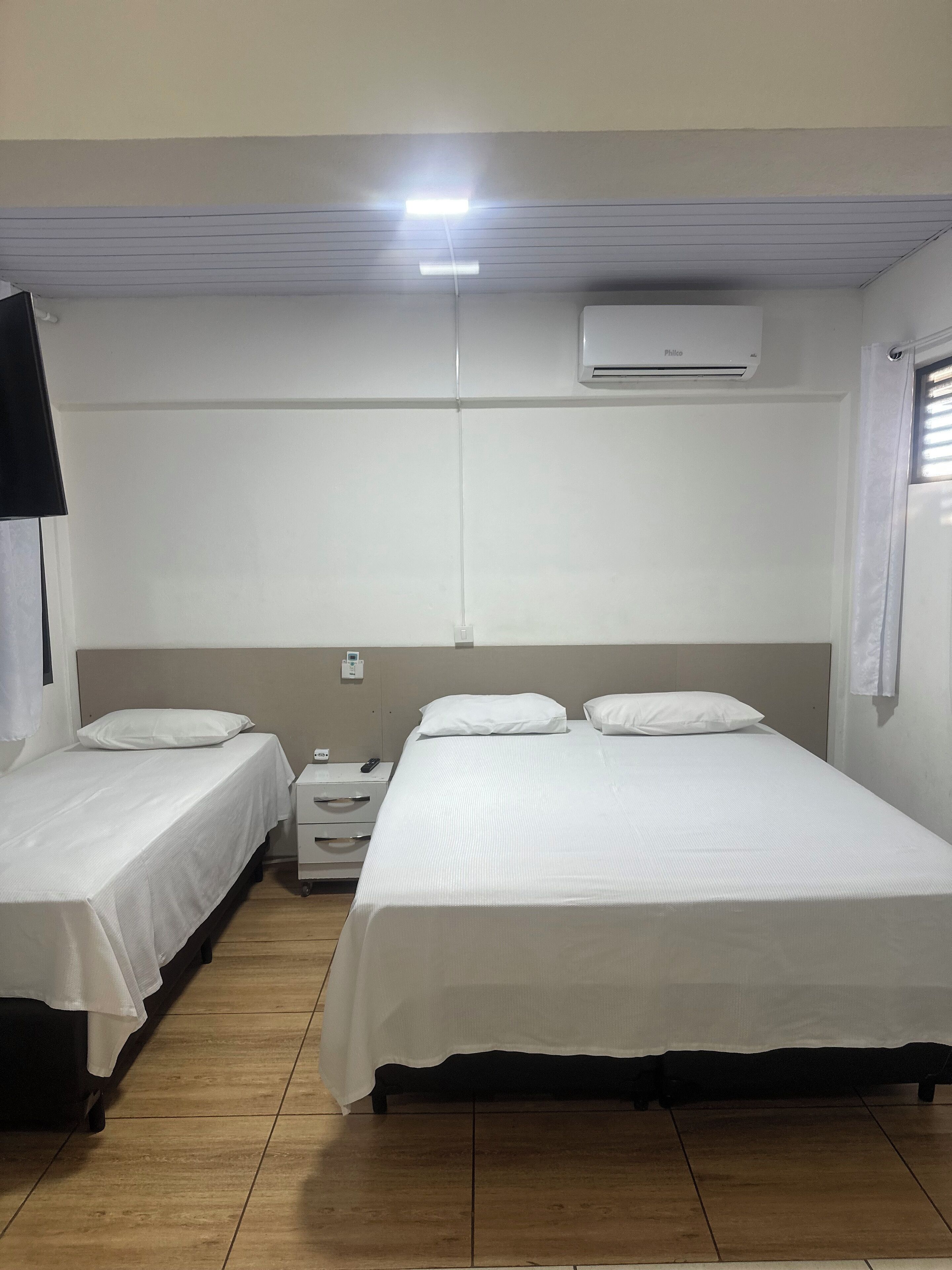 Quarto com duas camas de solteiro, mesa de cabeceira com abajur, ar condicionado fixado na parede e janela com persianas.