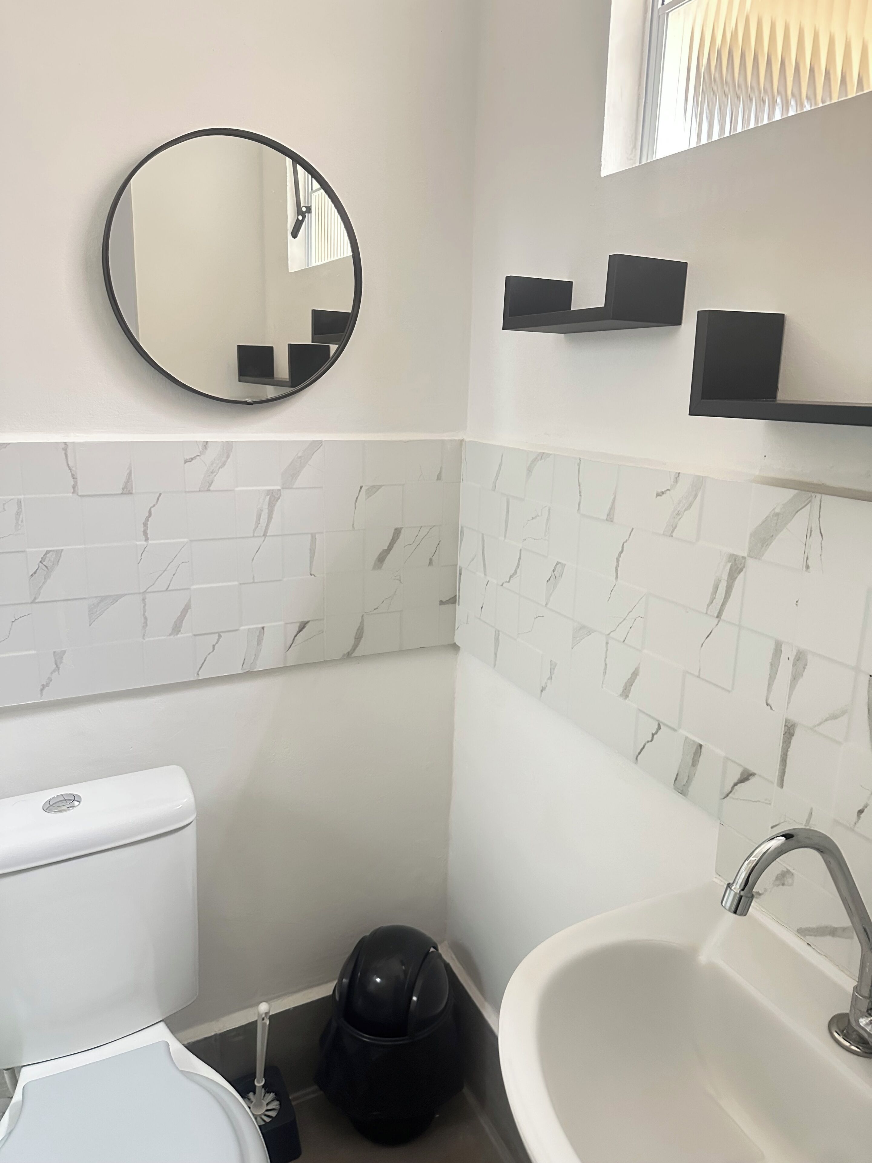A bathroom with a toilet, sink, mirror, and black storage shelf.