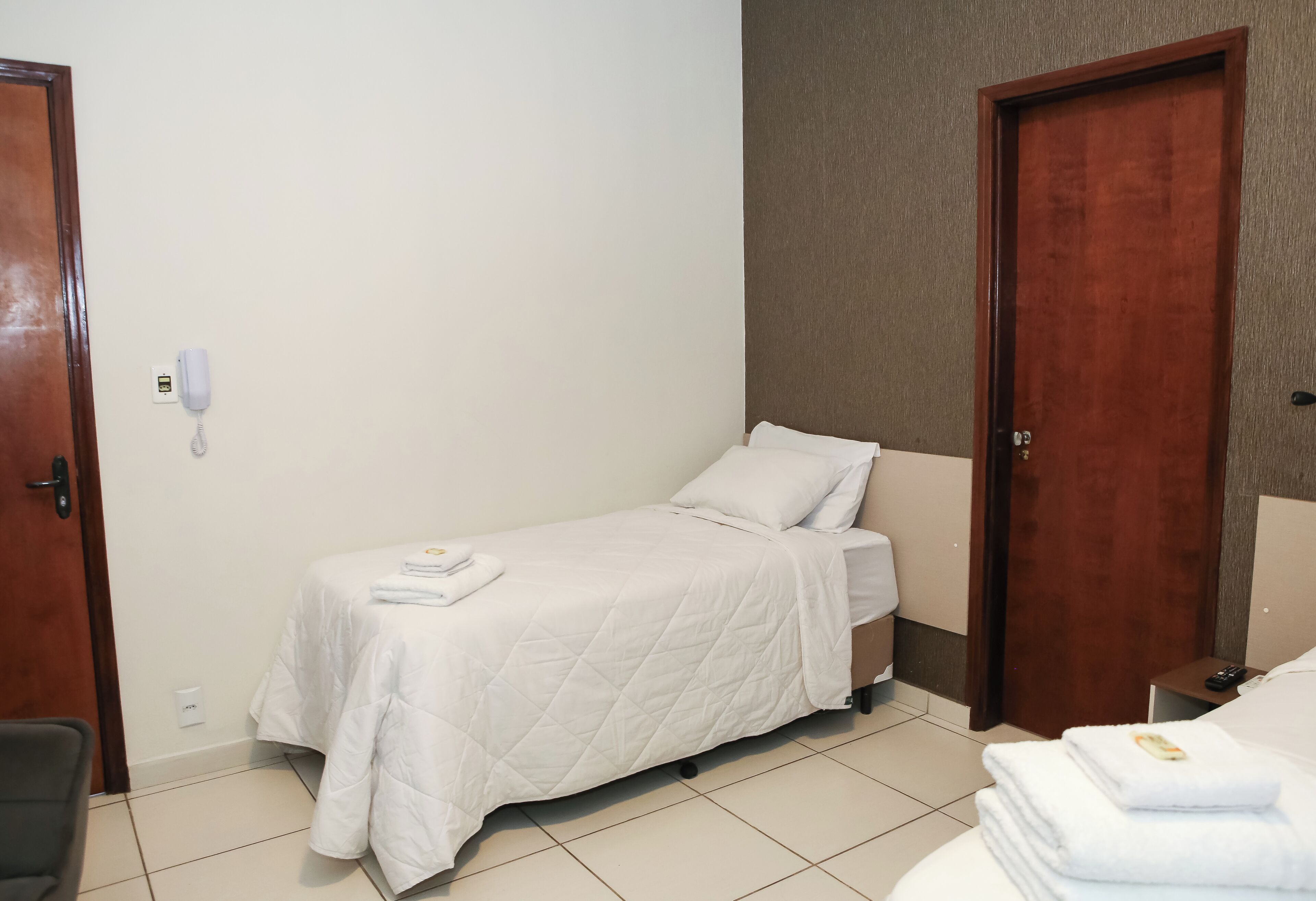 Una habitación pequeña con una cama individual, una colcha blanca, una mesita de noche, una silla y una puerta de madera.
