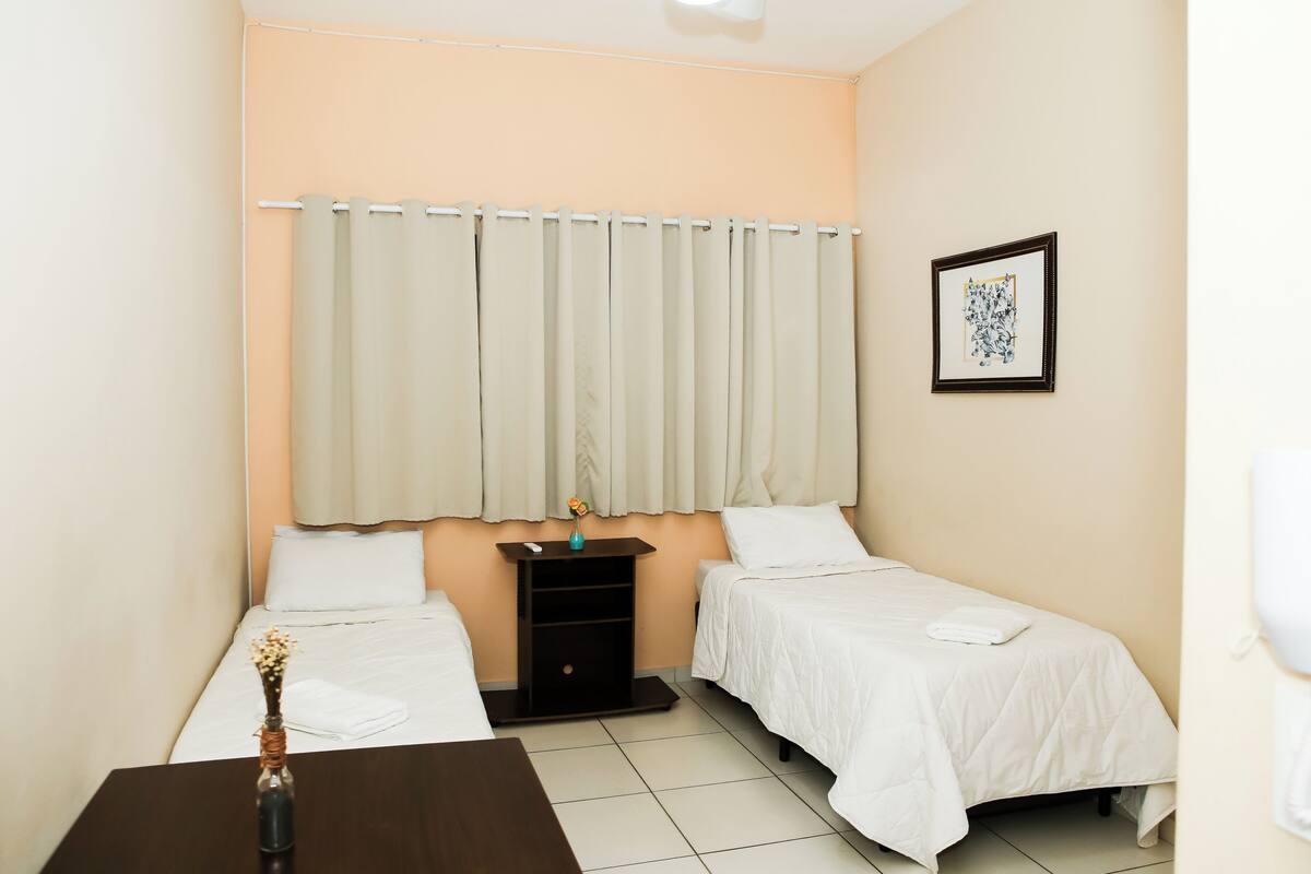 Quarto de hotel com duas camas, uma mesinha de cabeceira, uma pequena mesa, um vaso com flores e um quadro na parede.