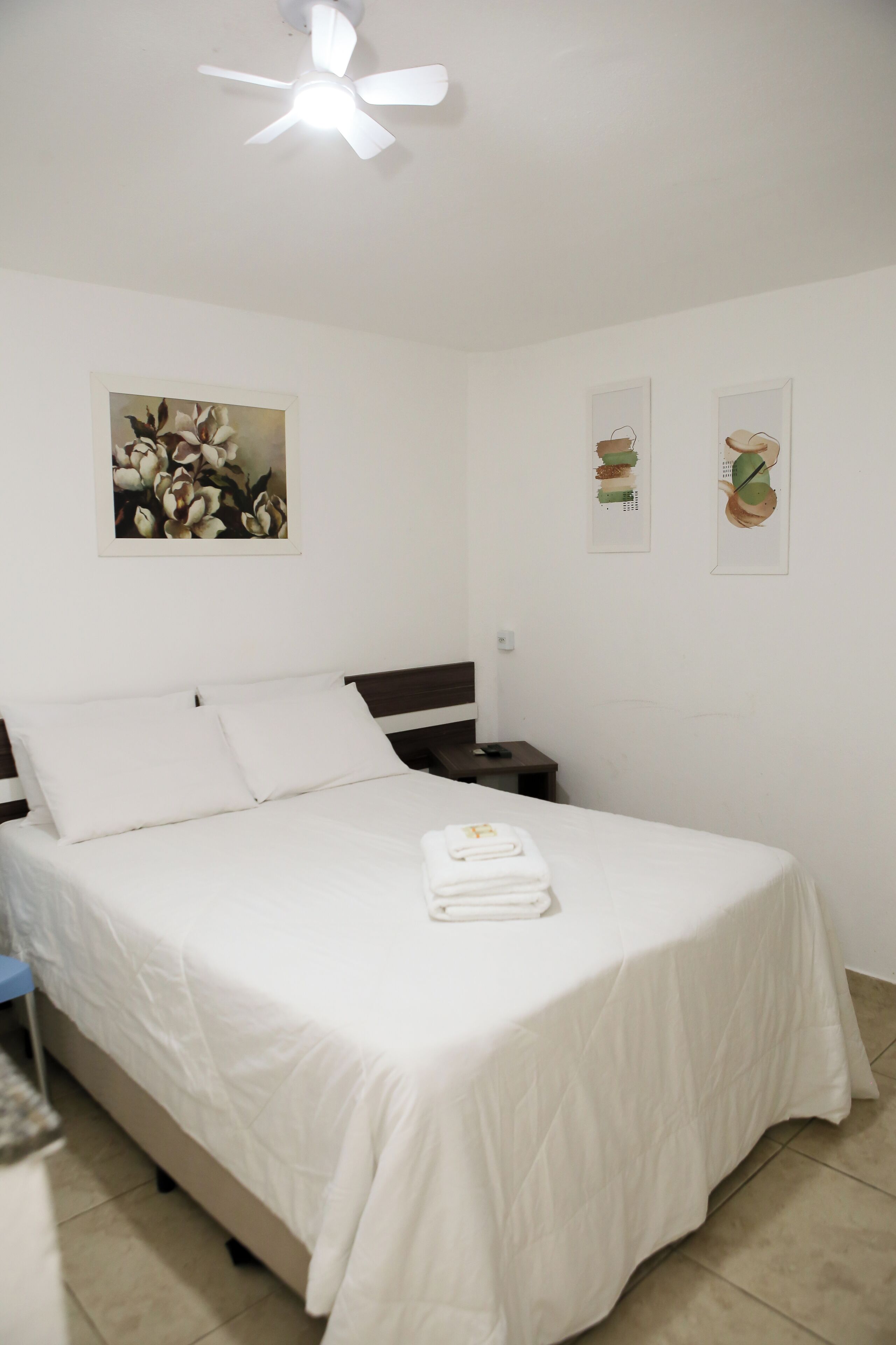 Een keurig opgemaakt bed met witte lakens, een nachtkastje met een lamp en een plafondventilator.