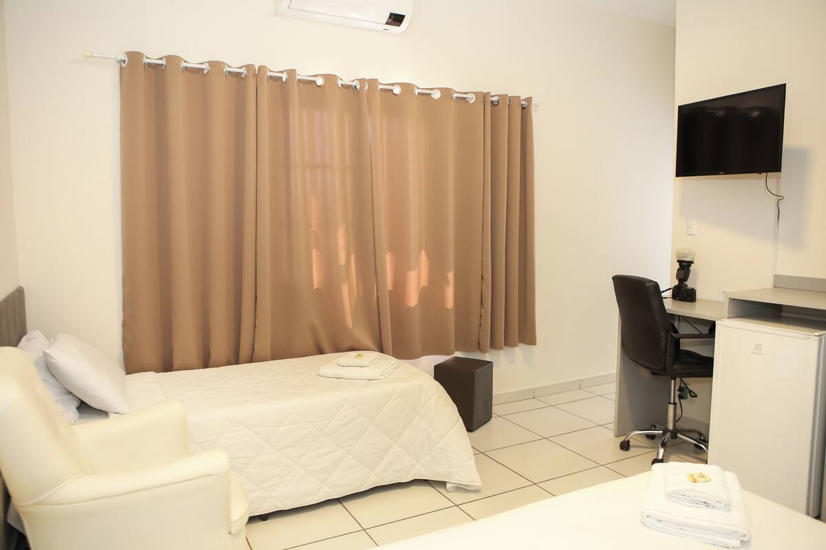 Quarto de hotel com cama, escrivaninha, cadeira e televisão.