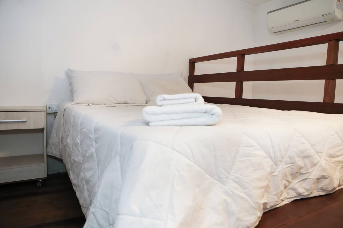 Uma cama bem arrumada com roupa de cama branca e cabeceira de madeira, uma mesinha de cabeceira com gaveta e um aparelho de ar condicionado fixado na parede.
