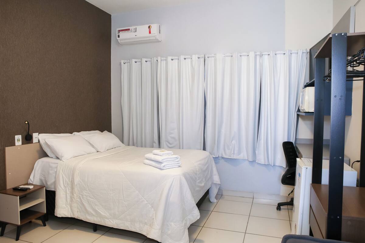 Quarto de hotel com cama, escrivaninha, cadeira, ar condicionado e cortina.