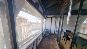 Un balcón estrecho con vistas a edificios, una mesa con sillas y un panel de vidrio.