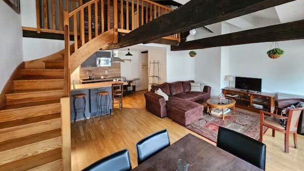 Amplia sala de estar con escalera de madera, un conjunto de sofás, una mesa de comedor y una zona de cocina.