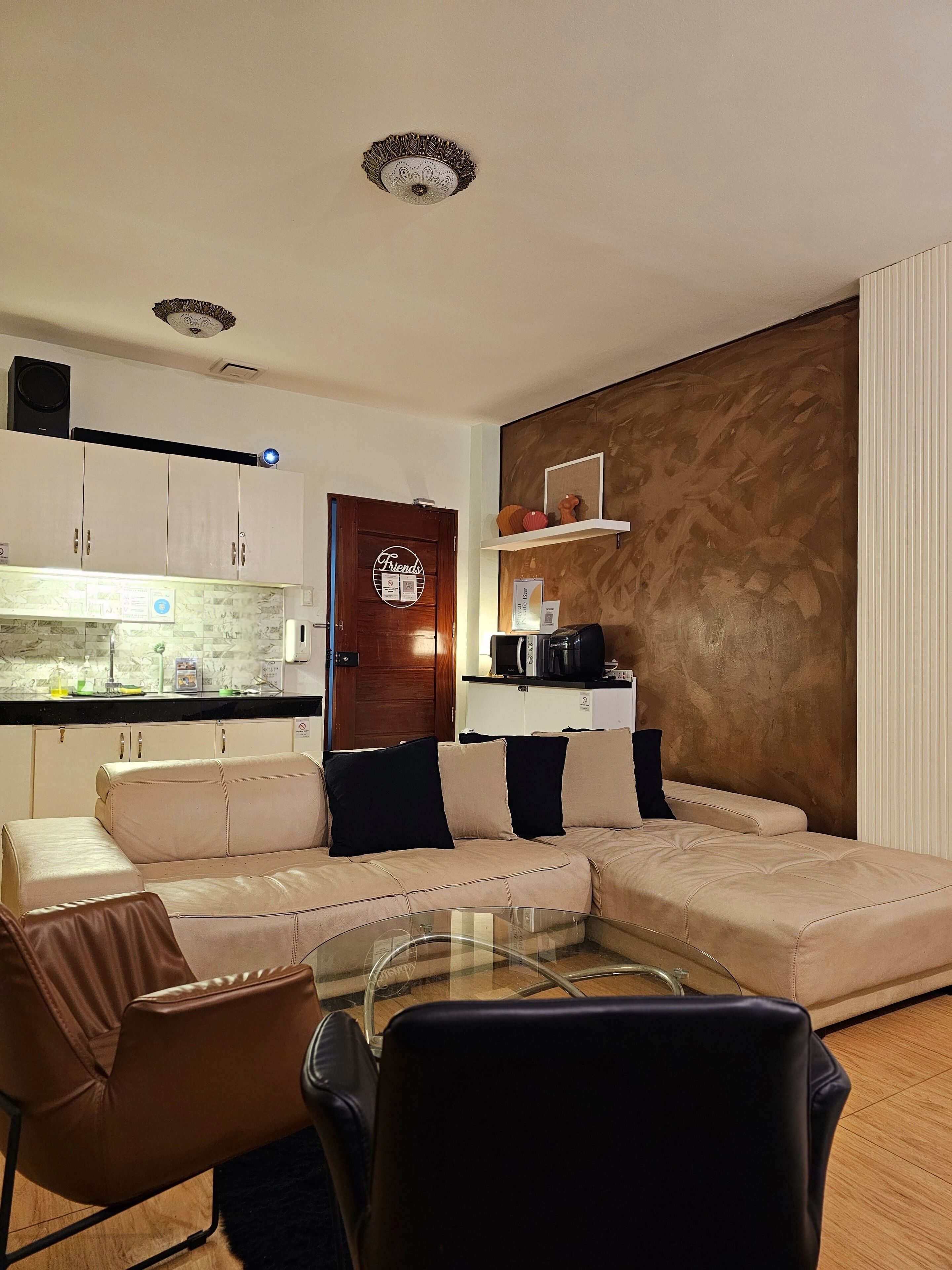 Ein modernes Wohnzimmer mit einer Couch, einem Glastisch und einem Essbereich mit Stühlen. Es gibt eine Küchenzeile mit Schränken und einer Mikrowelle.
