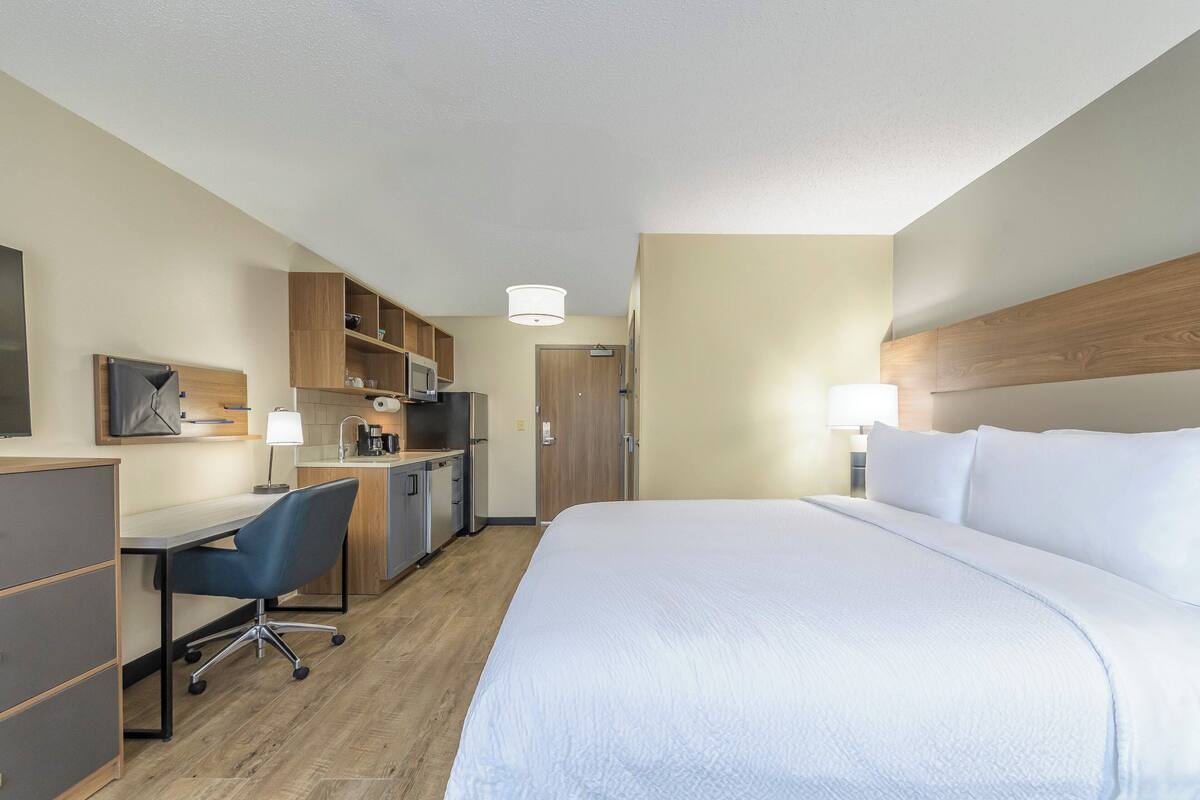 A hotel room with a bed, desk, chair, kitchenette, and a door.