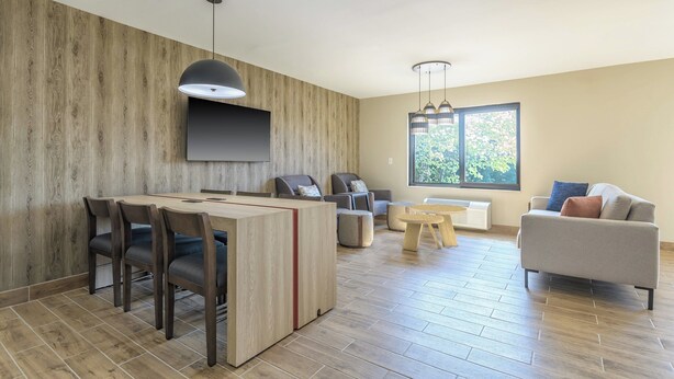 A modern living room with a wooden accent wall, a flat-screen TV, a dining area with chairs, a sofa, and a large window providing natural light.