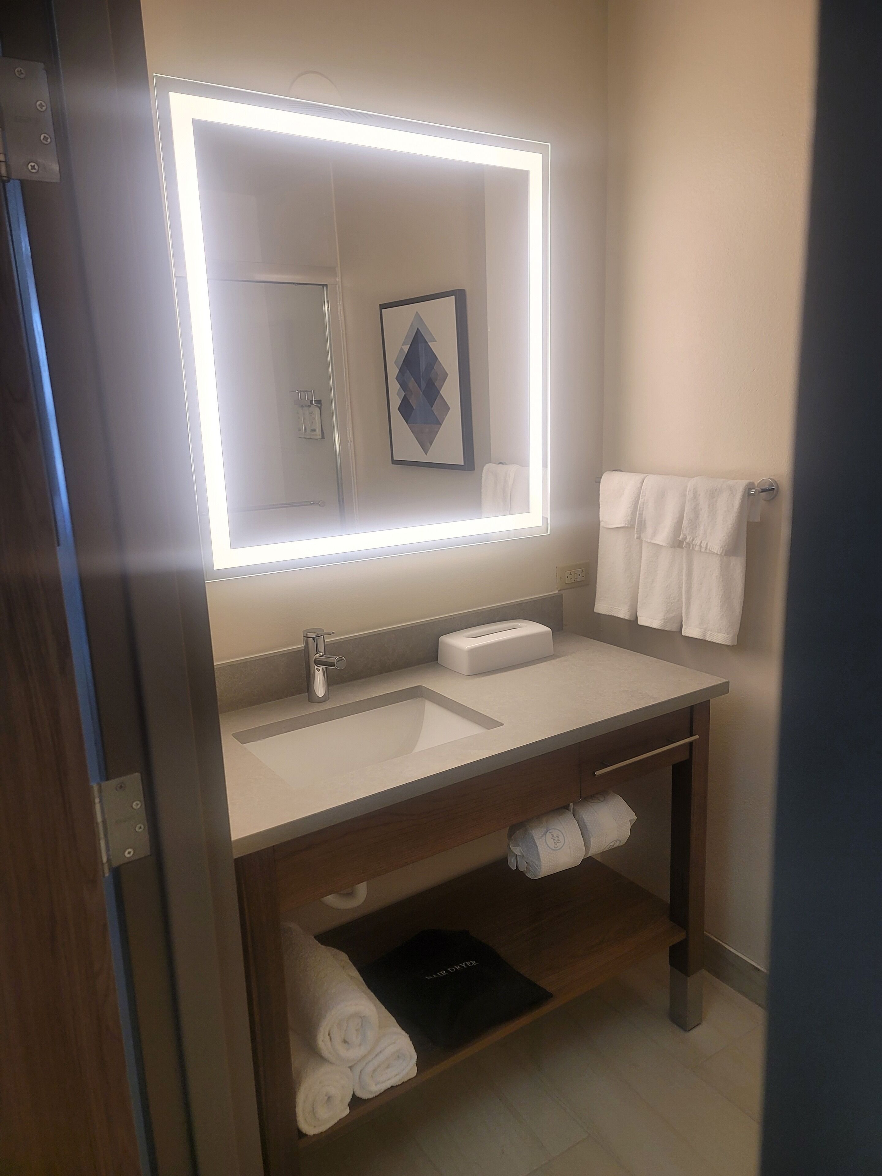 A modern bathroom with a large mirror, a sink, a towel rack, and a shelf with towels.