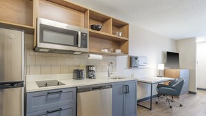 A modern kitchen with a built-in microwave, coffee maker, and dishwasher. There is a wooden shelf above the sink and a flat-screen TV mounted on the wall.