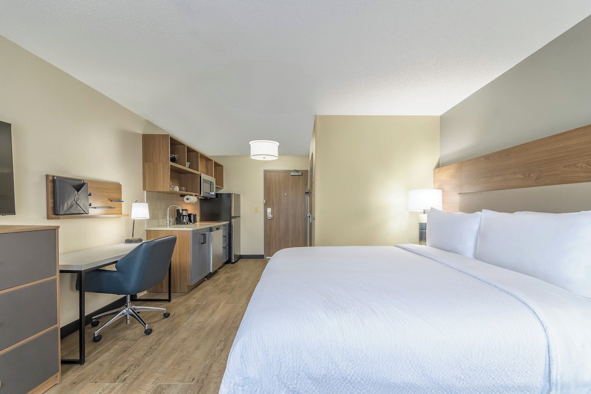A hotel room with a bed, desk, chair, kitchenette, and a door.