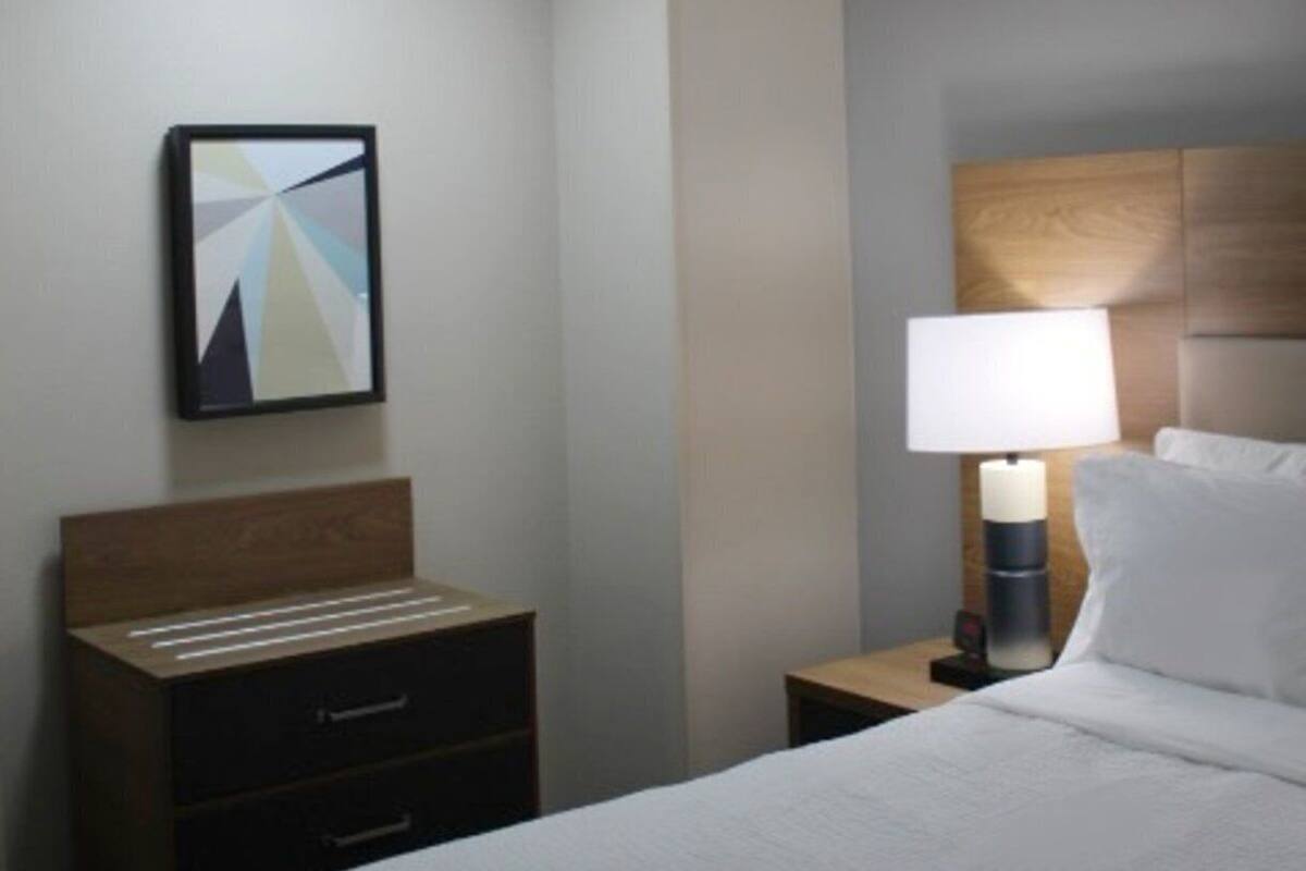 A hotel room with a bed, a bedside table with a lamp, a nightstand, a drawer unit, and a ceiling light.