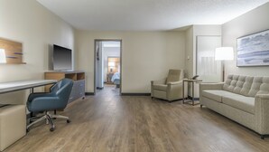 A hotel room with a desk, a blue office chair, a beige sofa, a lamp, and a television.