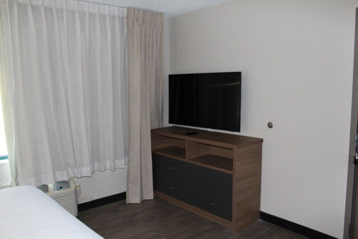 Quarto de hotel com uma cama, uma televisão fixada em um armário de madeira e uma janela com cortinas.