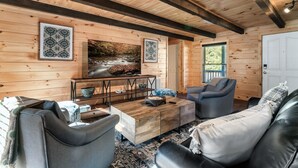 A cozy log cabin interior with a wooden ceiling, a large painting of a river, and a modern seating area with a coffee table. - River Access! Kayak/ Fire pit/ Hot Tub/ Top 10% (Sevierville)