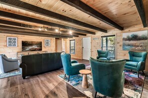 A modern living room with wooden walls and ceiling, a black sofa, teal armchairs, a wooden coffee table, and artwork on the walls.