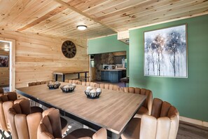 A spacious dining area with a long wooden table and leather chairs, a clock on the wall, and a kitchen in the background. - River Access! Kayak/ Fire pit/ Hot Tub/ Top 10% (Sevierville)