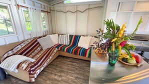 A cozy, well-lit room with a large, colorful sofa, a wooden table with a fruit basket, and a view of greenery outside. - Kukui Cabin with Private Bath (Kealakekua)